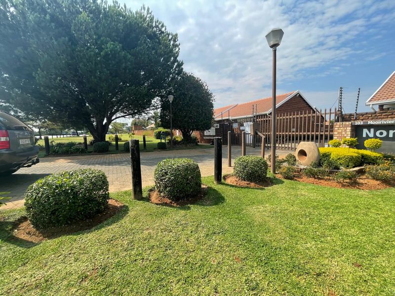 Front entrance view of the townhouse complex featuring a paved driveway, landscaped garden, and secure perimeter fencing.