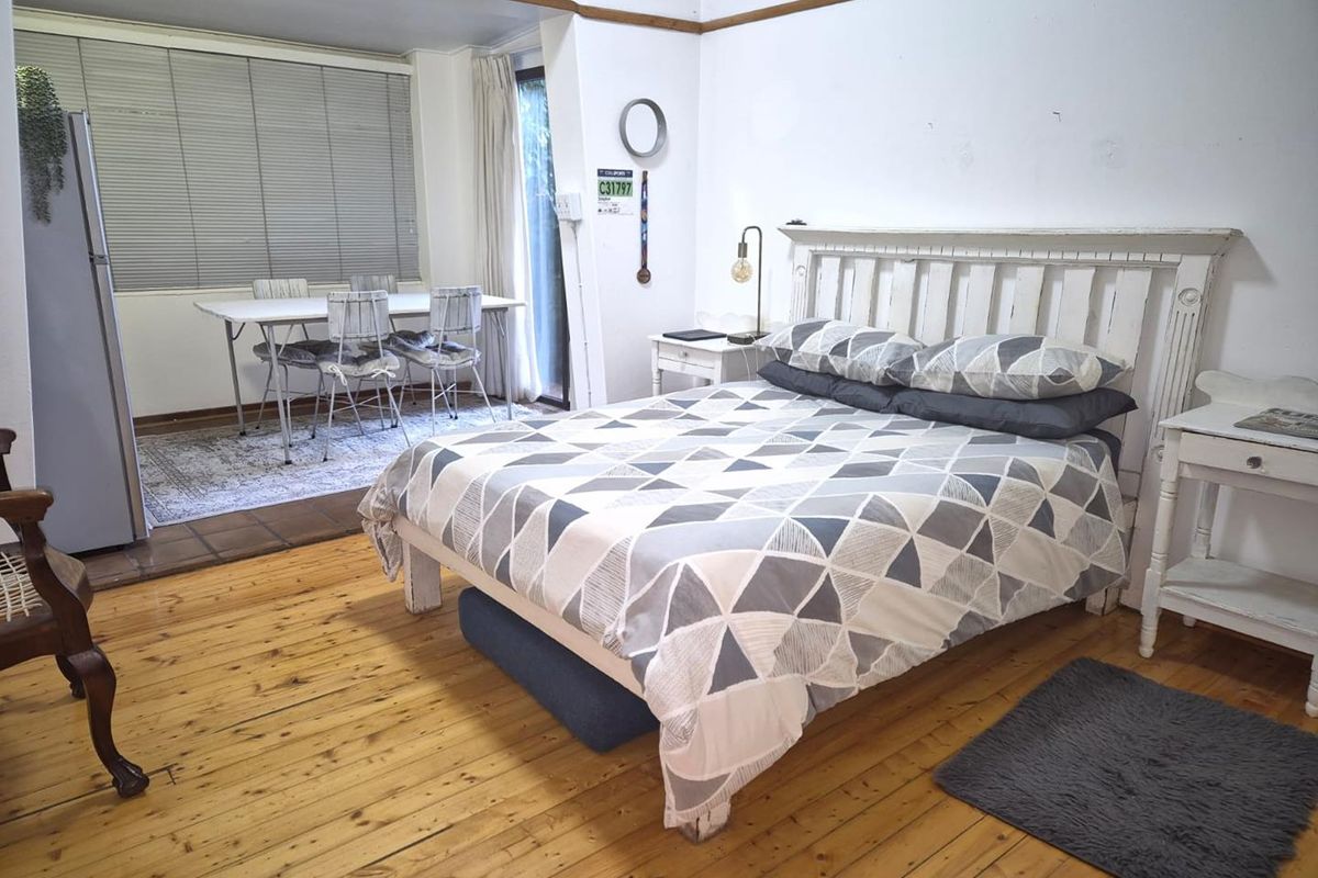 A spacious bedroom featuring polished timber flooring, neutral walls, and an adjoining dining area with large windows providing natural light.