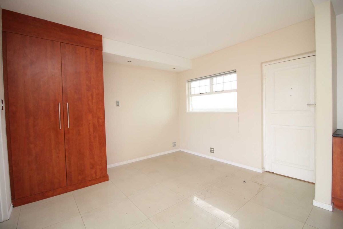 A bright, empty bedroom featuring neutral tiled flooring, a built-in wooden wardrobe, and recessed ceiling lighting.