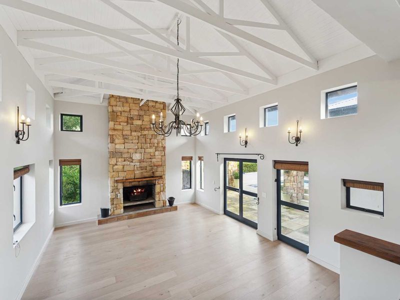 Spacious living room featuring a striking stone fireplace, high vaulted ceilings with exposed beams, hardwood floors, and multiple glass doors leading to the exterior.