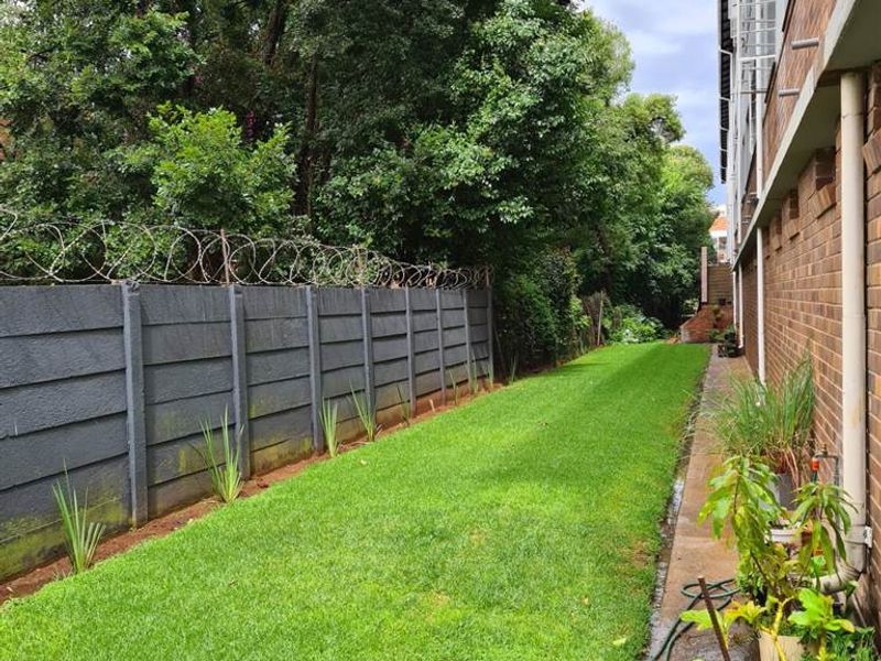 A side garden area featuring a well-maintained lawn, a brick building facade, and a secure boundary wall topped with razor wire.
