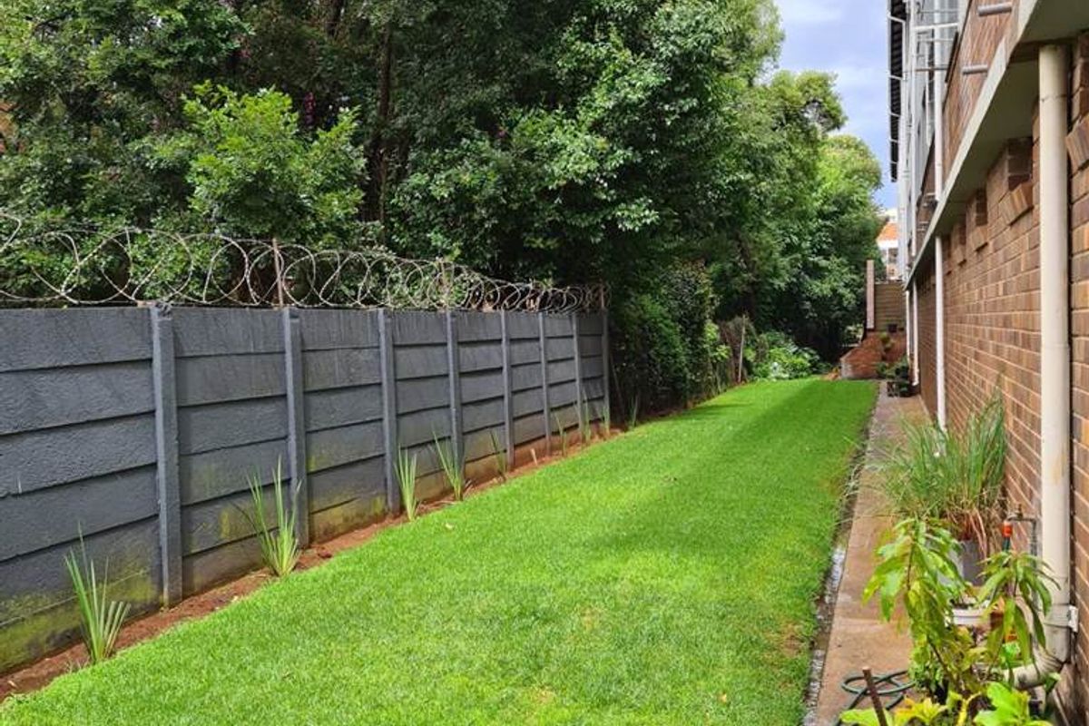A side garden area featuring a well-maintained lawn, a brick building facade, and a secure boundary wall topped with razor wire.