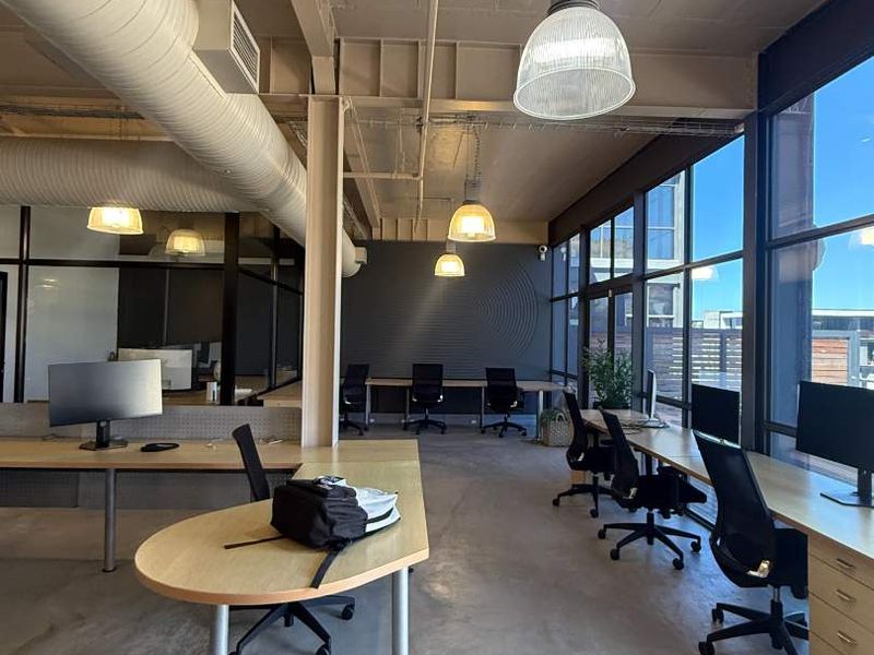 Modern industrial-style office space featuring exposed concrete ceilings, ductwork, polished concrete floors, and large windows providing ample natural light.