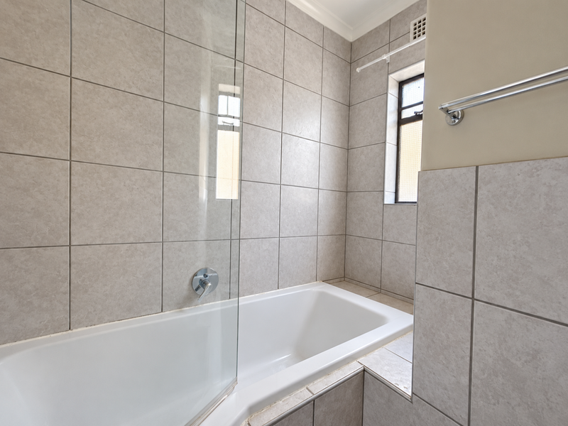 A clean bathroom featuring a built-in bathtub with a glass screen, neutral wall tiling, and chrome fixtures.