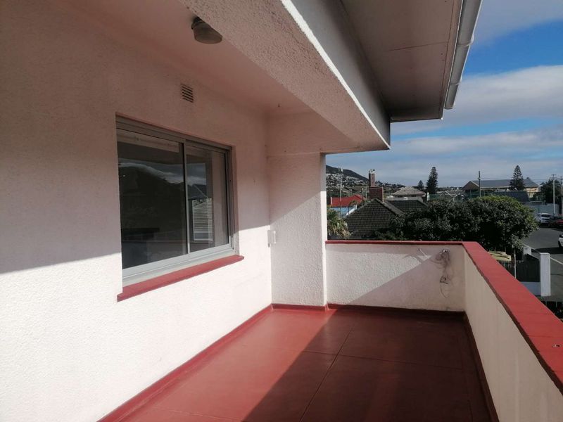 An open-air balcony featuring a painted concrete floor, parapet wall, and sliding window access, offering views of the surrounding neighborhood.