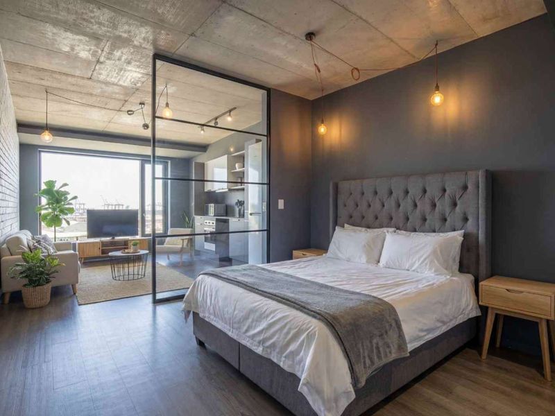Modern open-plan apartment featuring an exposed concrete ceiling, glass partition separating the bedroom from the living area, and a sleek integrated kitchen.