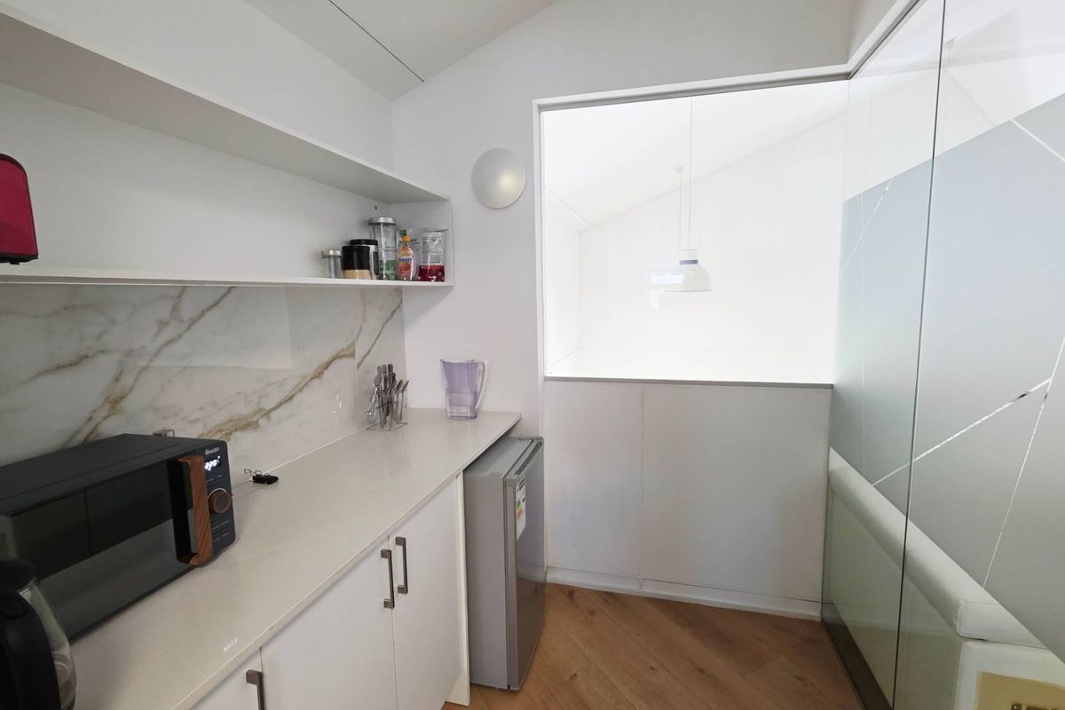 Modern kitchenette featuring marble-effect splashback, sleek white cabinetry, stone countertops, and a glass partition wall, set against warm timber flooring.
