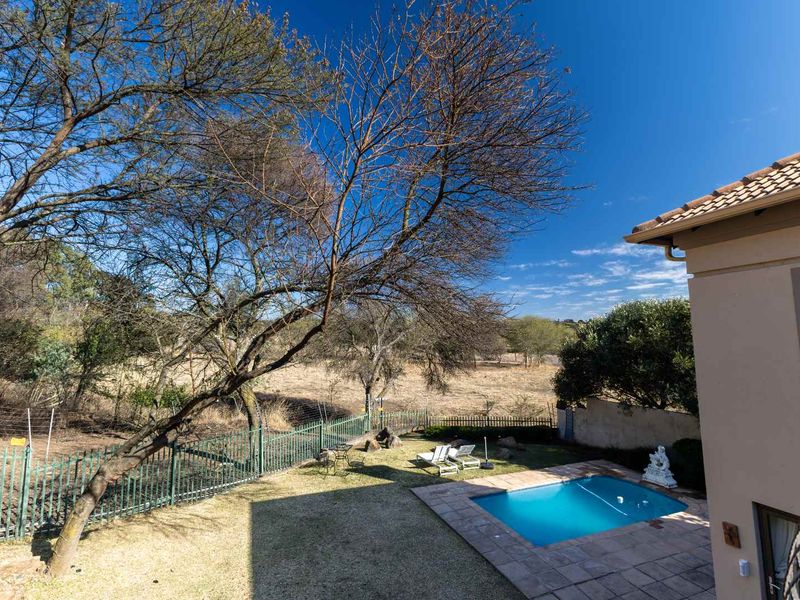 A view of the rear garden featuring a swimming pool with tiled surround, metal fencing, and mature trees overlooking an open field.