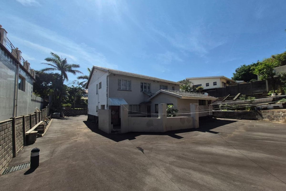 Rear view of the property featuring a large asphalt driveway, perimeter walling, and a multi-level structure with tiled roofing.