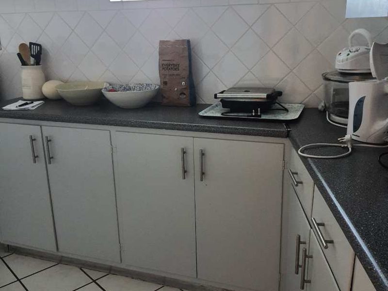 A functional kitchen featuring laminate countertops, tiled backsplash, and ample cabinetry, with ceramic floor tiles and natural light from louvre windows.