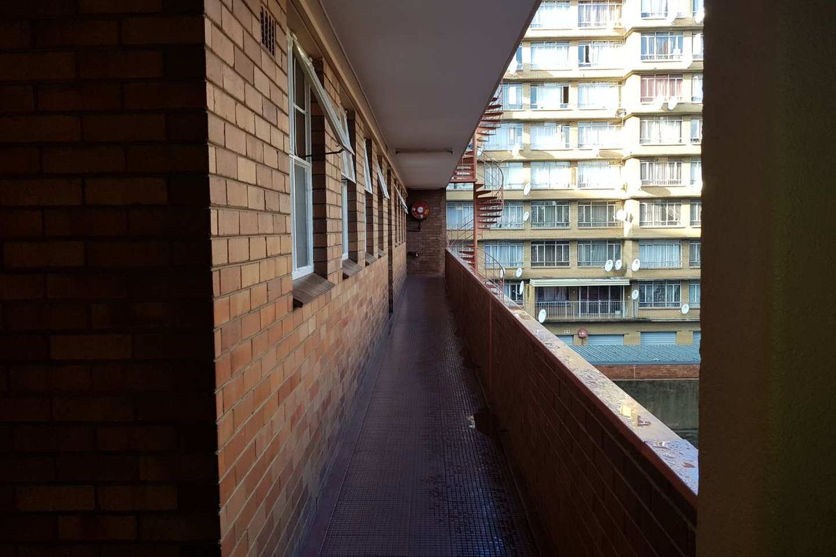An exterior walkway or balcony featuring brick walls and tiled flooring, providing access to the apartment units.