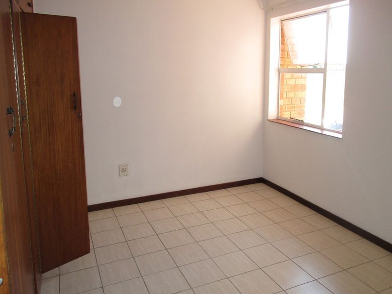 A clean, empty bedroom featuring neutral tiled flooring, built-in wooden cupboards, and a window providing natural light.