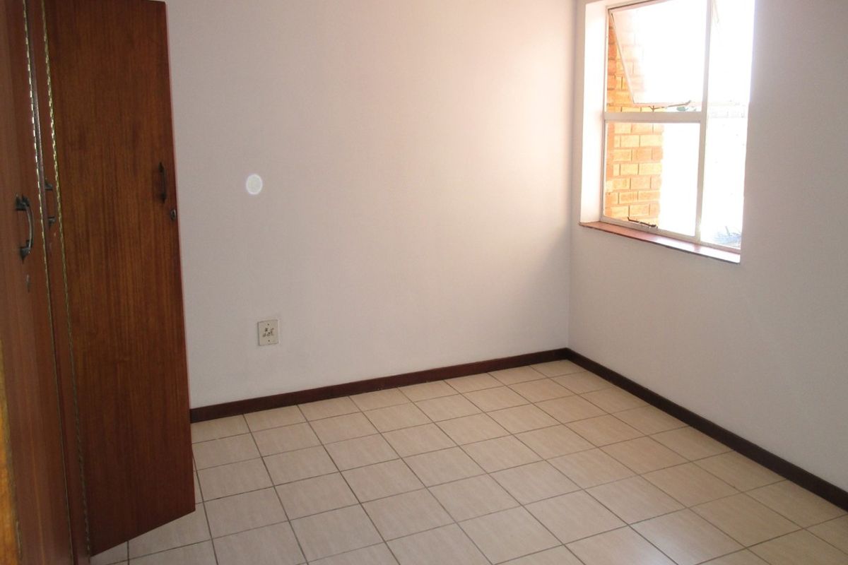 A clean, empty bedroom featuring neutral tiled flooring, built-in wooden cupboards, and a window providing natural light.