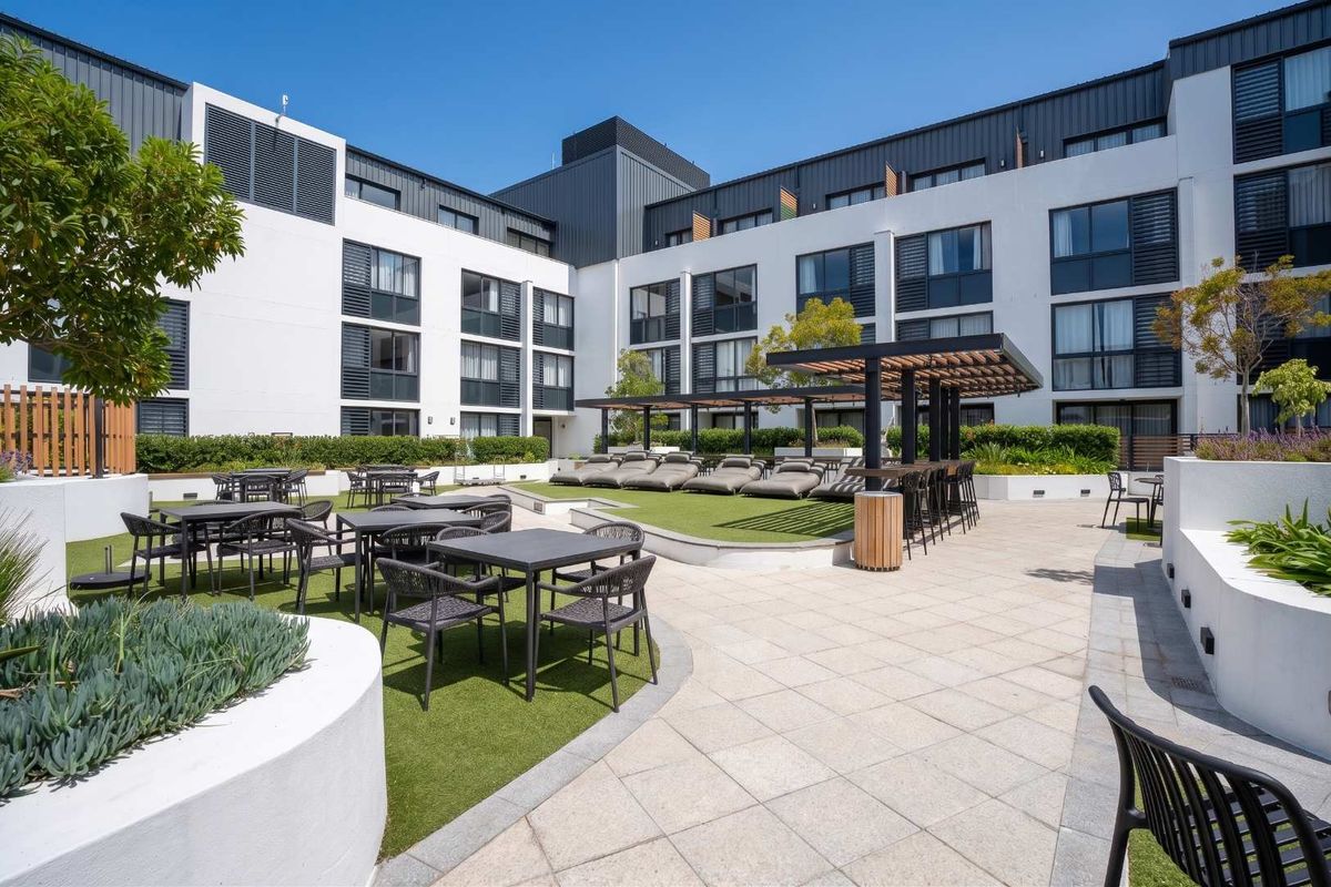 A well-maintained communal outdoor terrace featuring paved areas, artificial lawn, and a modern pergola structure within an apartment complex courtyard.