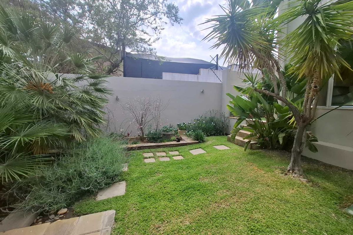 A private rear garden featuring a manicured lawn, stone stepping stones, established tropical landscaping, and a high perimeter boundary wall.