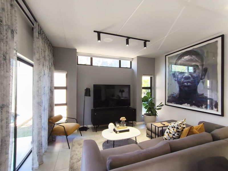 Modern living room featuring high ceilings with track lighting, large format floor tiles, and expansive windows providing natural light and garden access.