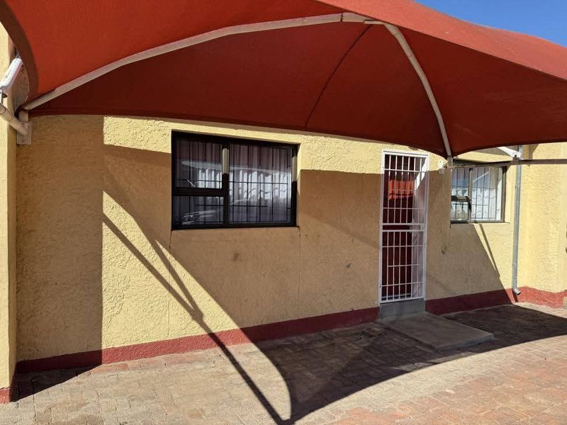 A paved outdoor patio area featuring a large red shade sail, security gate, and security bars on the windows.
