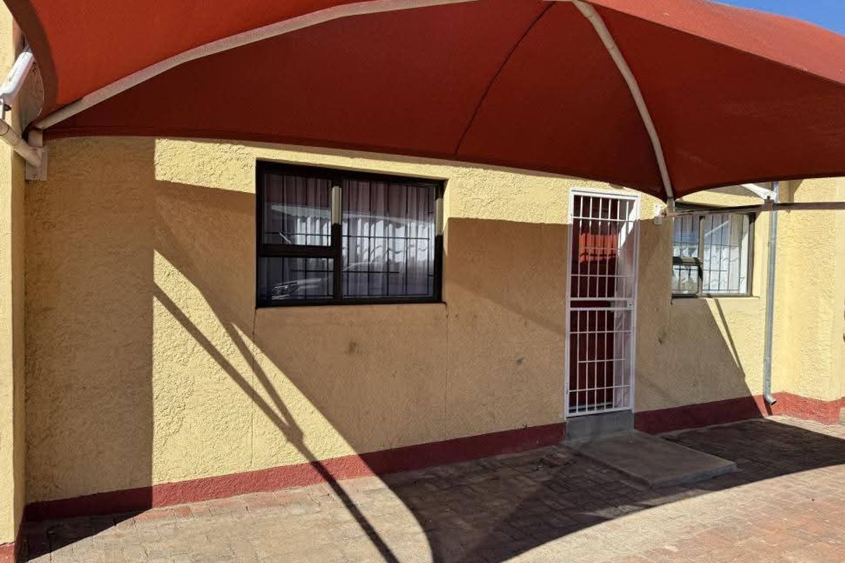 A paved outdoor patio area featuring a large red shade sail, security gate, and security bars on the windows.