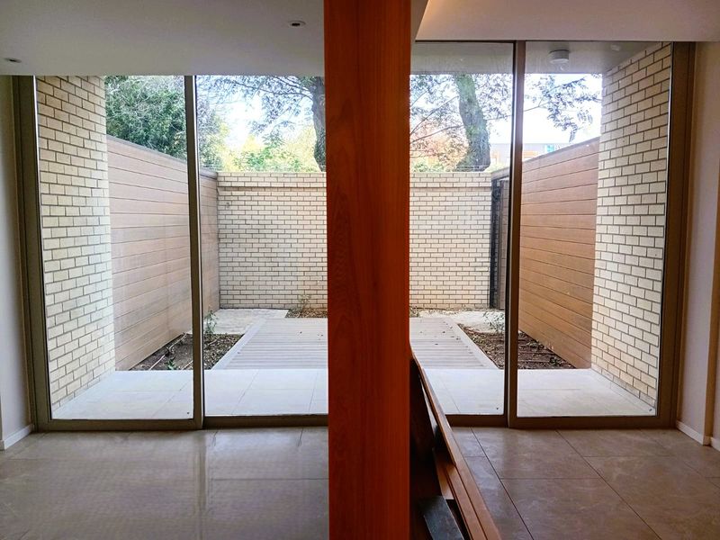 A modern, minimalist patio area featuring timber decking, brick boundary walls, and large sliding glass doors that provide seamless indoor-outdoor flow.