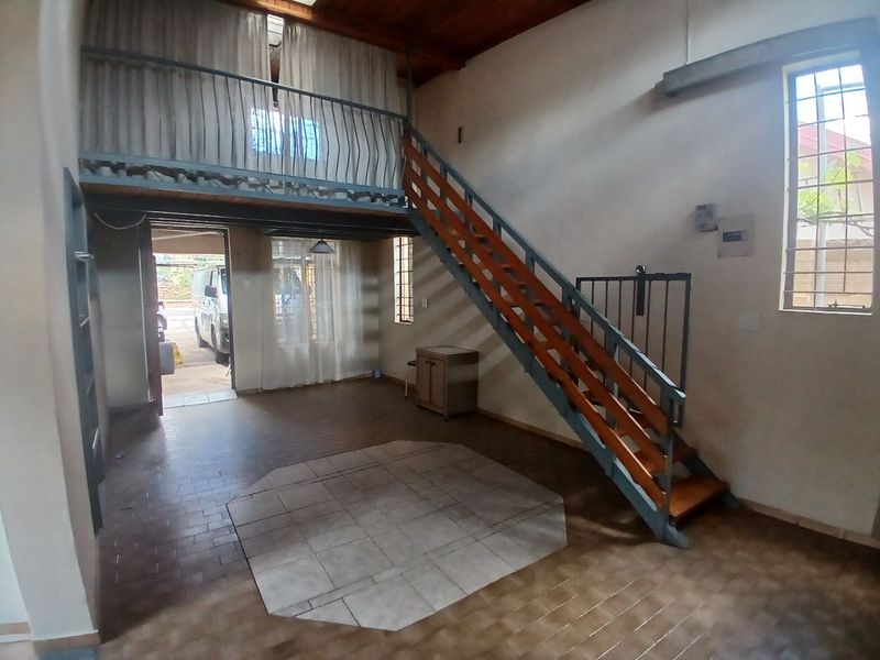Open-plan living area featuring a mezzanine level, timber staircase, exposed ceiling beams, and tiled flooring with security bars on windows.