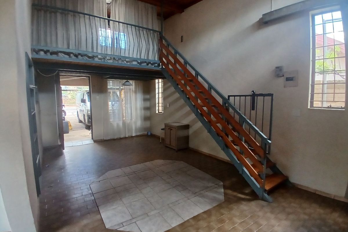 Open-plan living area featuring a mezzanine level, timber staircase, exposed ceiling beams, and tiled flooring with security bars on windows.