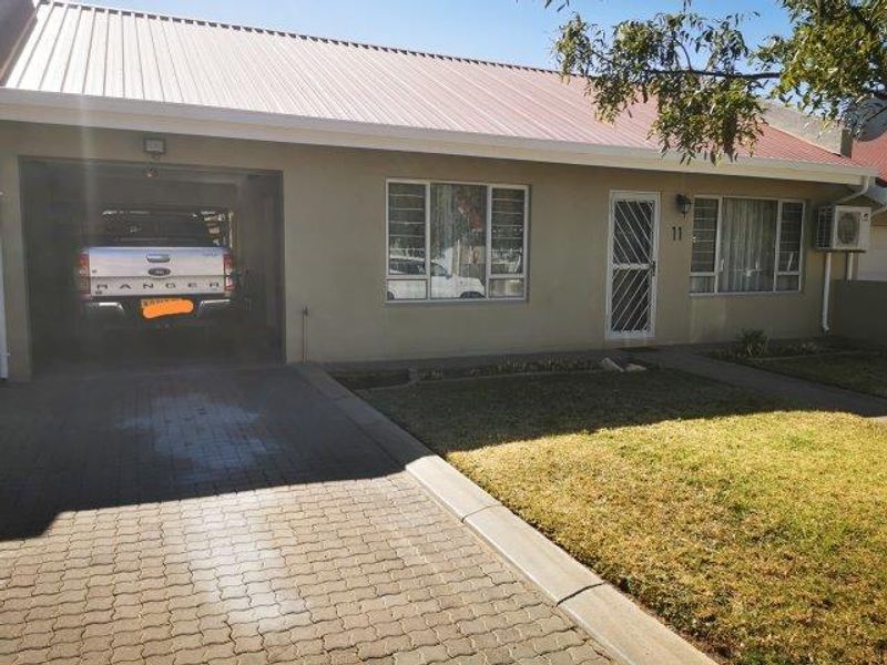 Front elevation of a townhouse featuring a paved driveway, integrated garage, metal roof, and security features including burglar bars and a security gate.