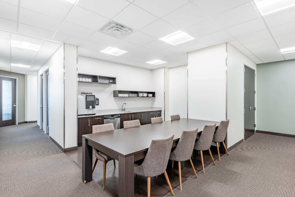 A professional office breakroom featuring a kitchenette with built-in cabinetry, sink, and a central meeting table with seating.