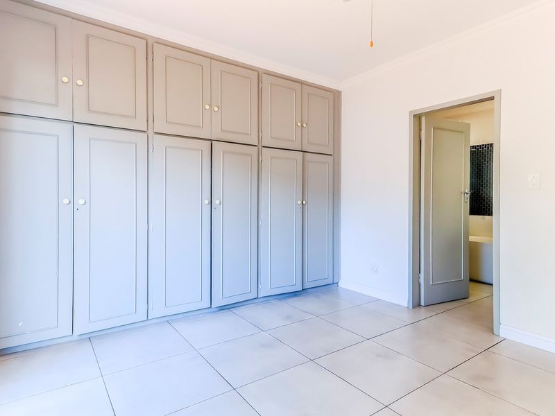 A bright, empty bedroom featuring extensive built-in cupboards, neutral tiled flooring, and a doorway leading to an adjacent bathroom.