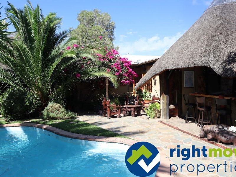 Outdoor pool area featuring a thatched roof entertainment space with a built-in bar and stone paved patio surrounded by established garden landscaping.