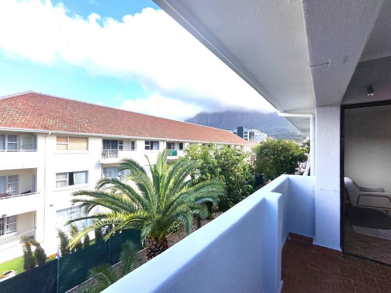A covered balcony featuring tiled flooring and a solid balustrade, offering an elevated view of the surrounding apartment complex and mountain landscape.