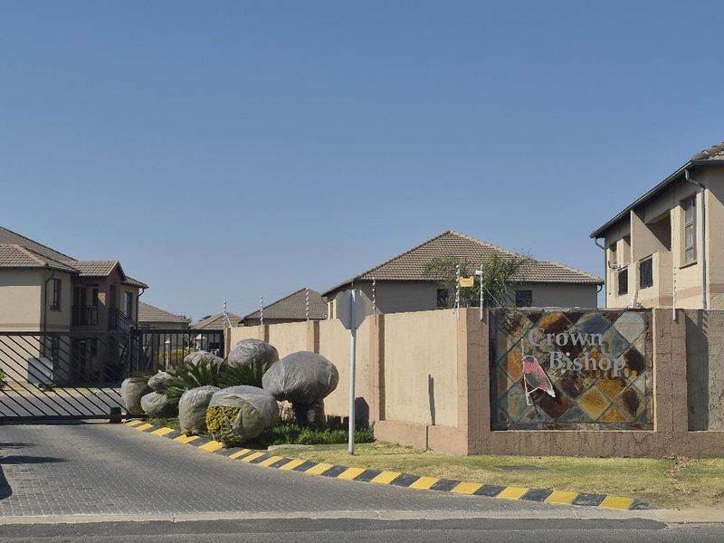 Front entrance view of the Crown Bishop apartment complex, featuring a paved driveway, security gate, and perimeter boundary wall with electric fencing.