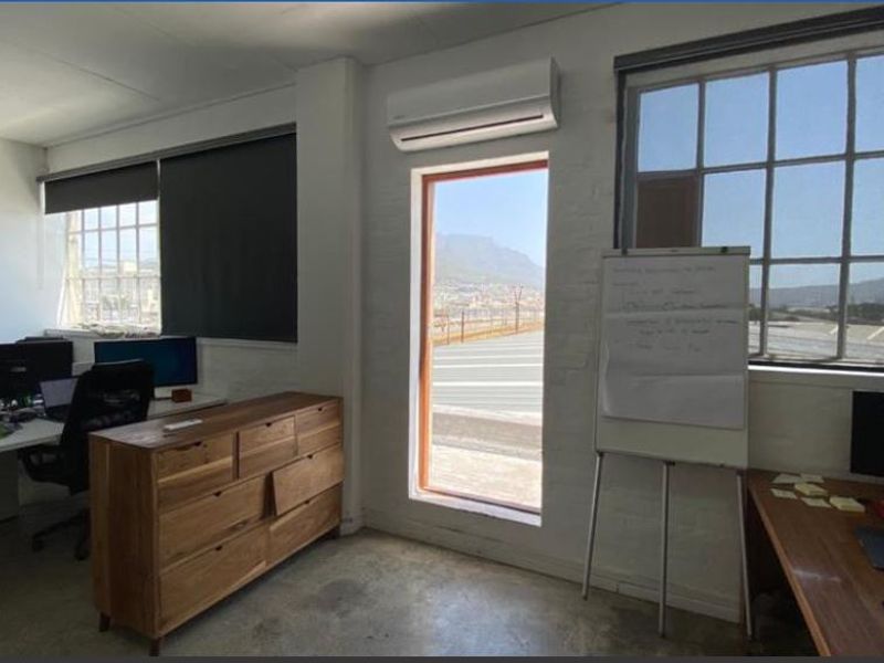 A bright office space featuring industrial-style windows, concrete flooring, and a wall-mounted air conditioning unit with views of the surrounding area.