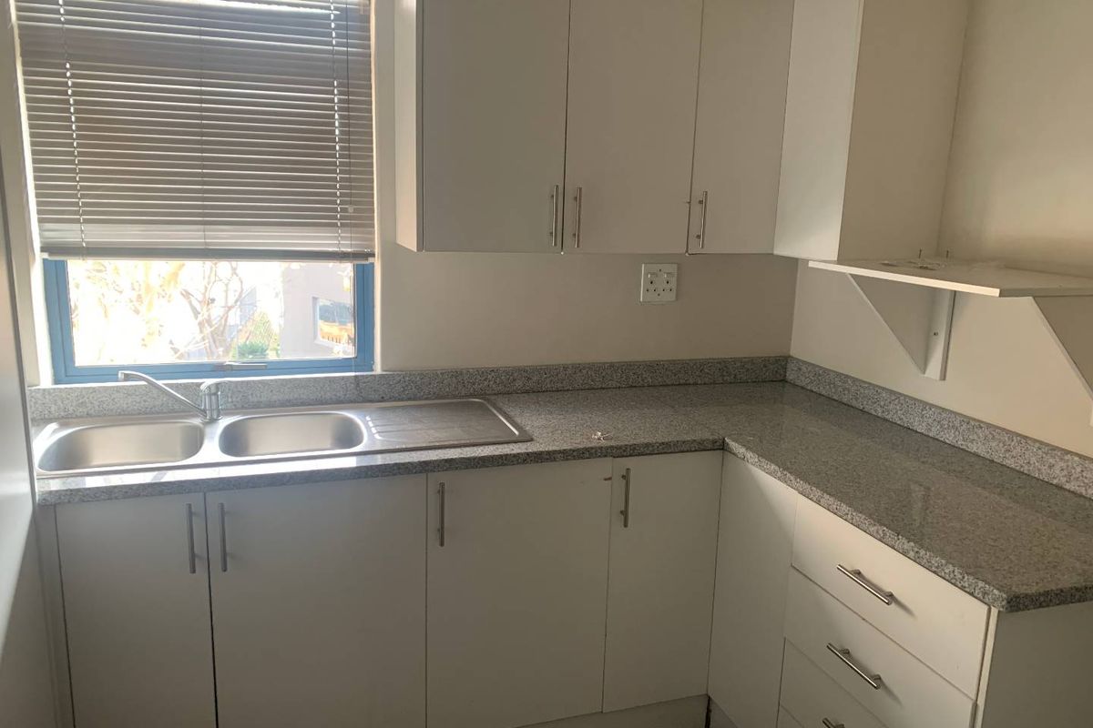 A functional office kitchenette featuring white cabinetry, granite-look countertops, a stainless steel double sink, and a window with fitted blinds.