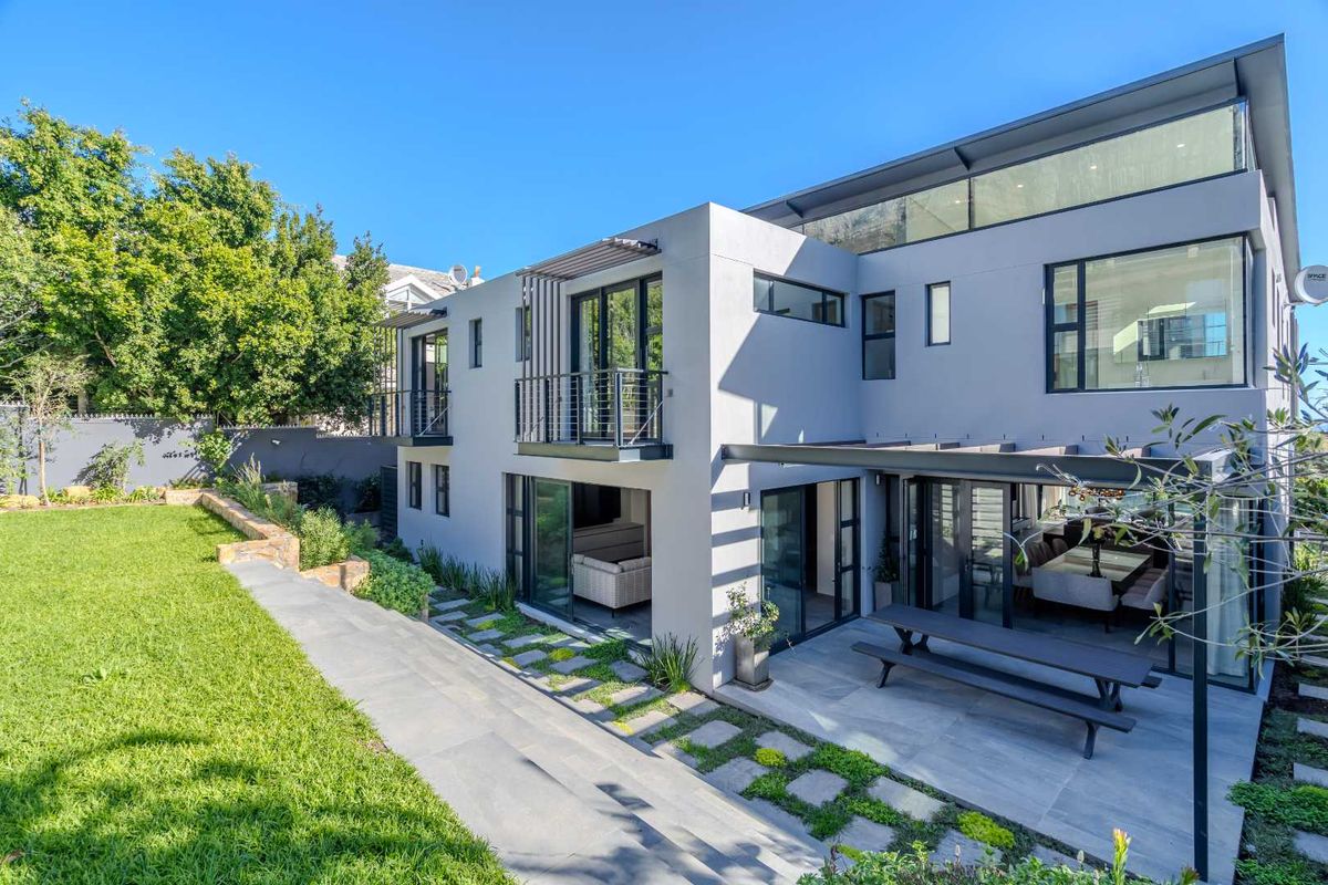 A modern rear exterior view of the house featuring a covered patio, landscaped garden, stone pathways, and large glass doors connecting indoor and outdoor spaces.