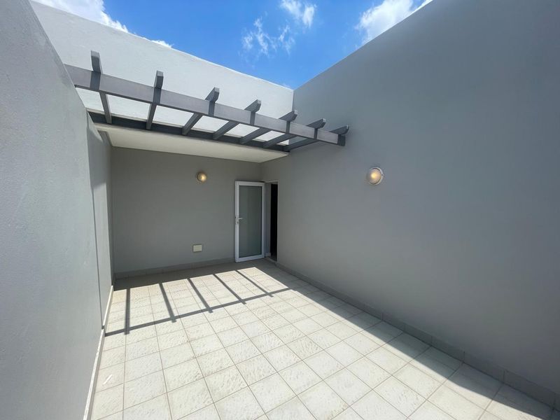 A clean, modern outdoor patio area featuring a pergola, tiled flooring, and wall-mounted lighting, providing a versatile space for outdoor relaxation.