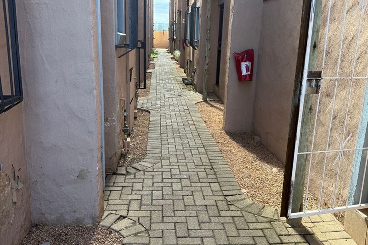 A narrow paved exterior walkway between apartment building sections featuring security gates and wall-mounted fire safety equipment.