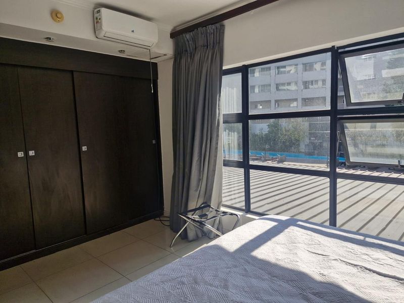 A bright bedroom featuring tiled flooring, dark built-in cupboards, a wall-mounted air conditioner, and large windows providing natural light.