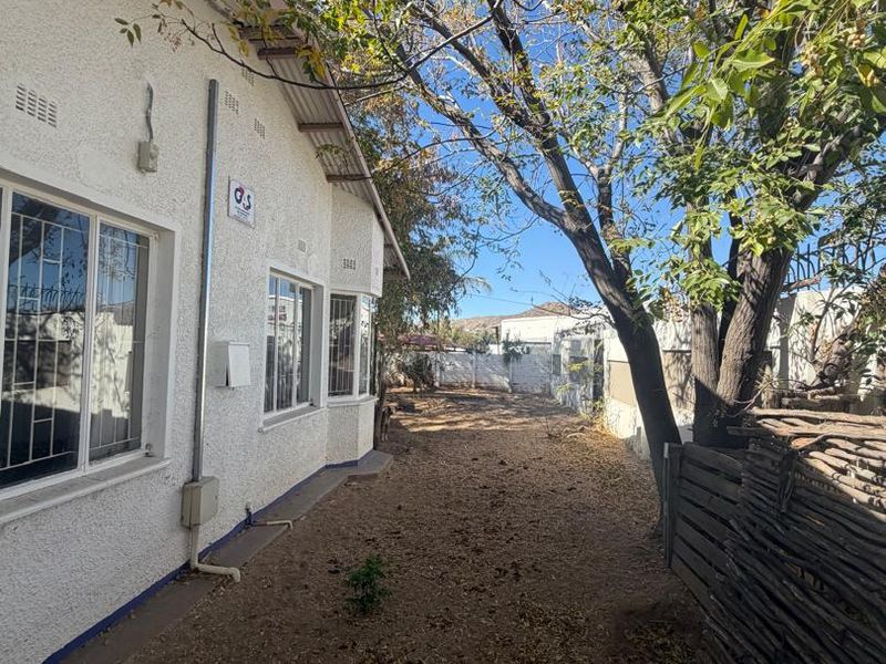 A view of the rear exterior side yard featuring a stucco facade, security bars on windows, and a large mature tree.