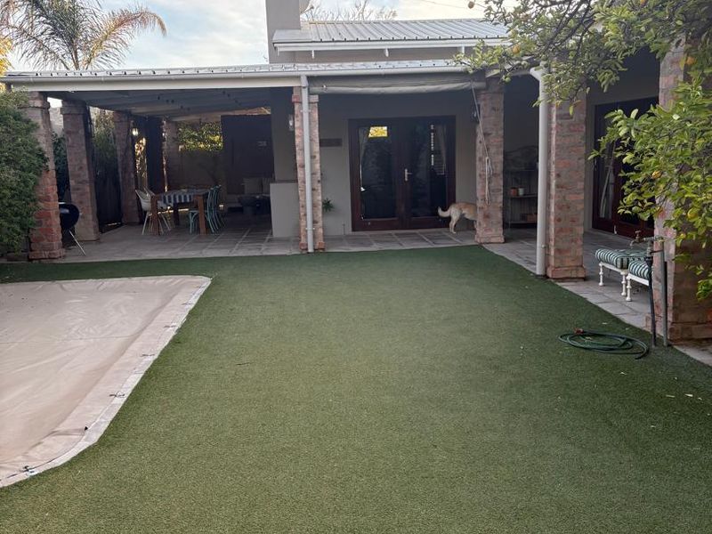 Rear exterior view featuring a covered patio with brick pillars, artificial lawn, and a swimming pool with a protective cover.