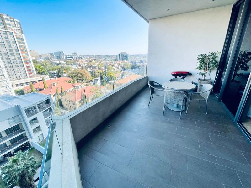 A spacious covered balcony featuring modern tiled flooring, a sleek glass balustrade, and expansive elevated city views.