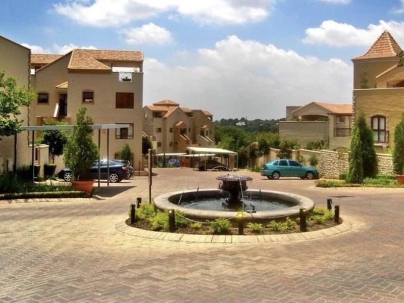 A view of the apartment complex entrance featuring a central fountain, paved driveway, and covered parking areas.