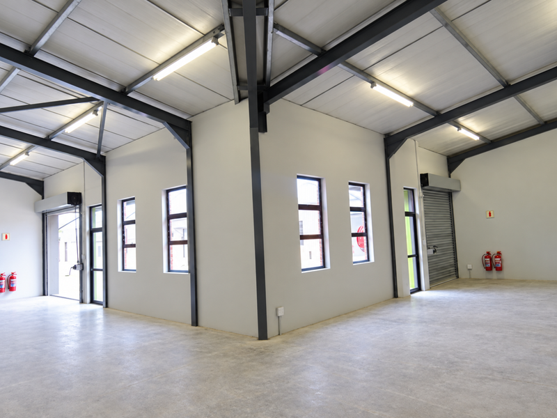 Spacious industrial utility area featuring high ceilings with exposed steel beams, concrete flooring, roller shutter doors, and functional fluorescent lighting.
