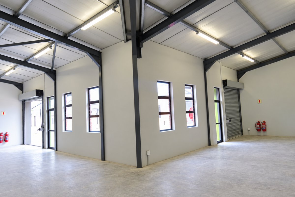 Spacious industrial utility area featuring high ceilings with exposed steel beams, concrete flooring, roller shutter doors, and functional fluorescent lighting.