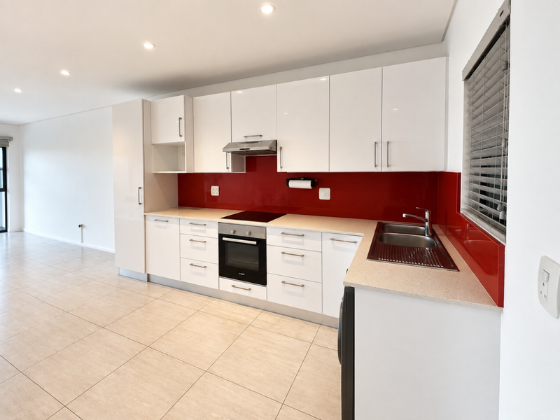 Modern open-plan kitchen featuring white cabinetry, a striking red glass splashback, integrated appliances, and neutral tiled flooring in a bright, spacious living area.