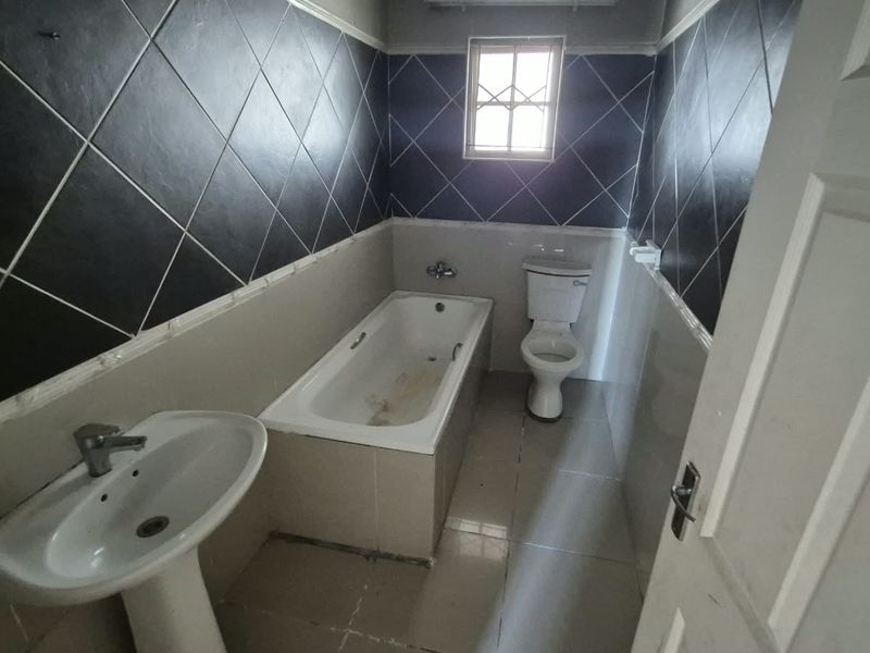 A dated bathroom featuring a pedestal basin, built-in bathtub, and toilet, with dark diamond-patterned wall tiles and neutral floor tiling.
