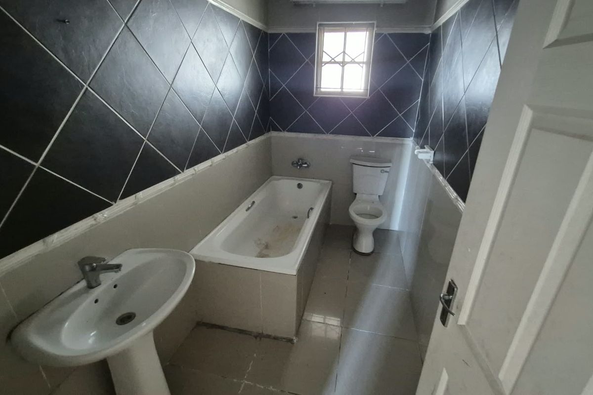 A dated bathroom featuring a pedestal basin, built-in bathtub, and toilet, with dark diamond-patterned wall tiles and neutral floor tiling.