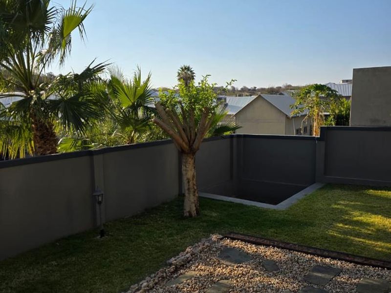 A well-maintained private garden featuring a stone-paved area, manicured lawn, and a modern boundary wall with a unique sunken architectural feature.