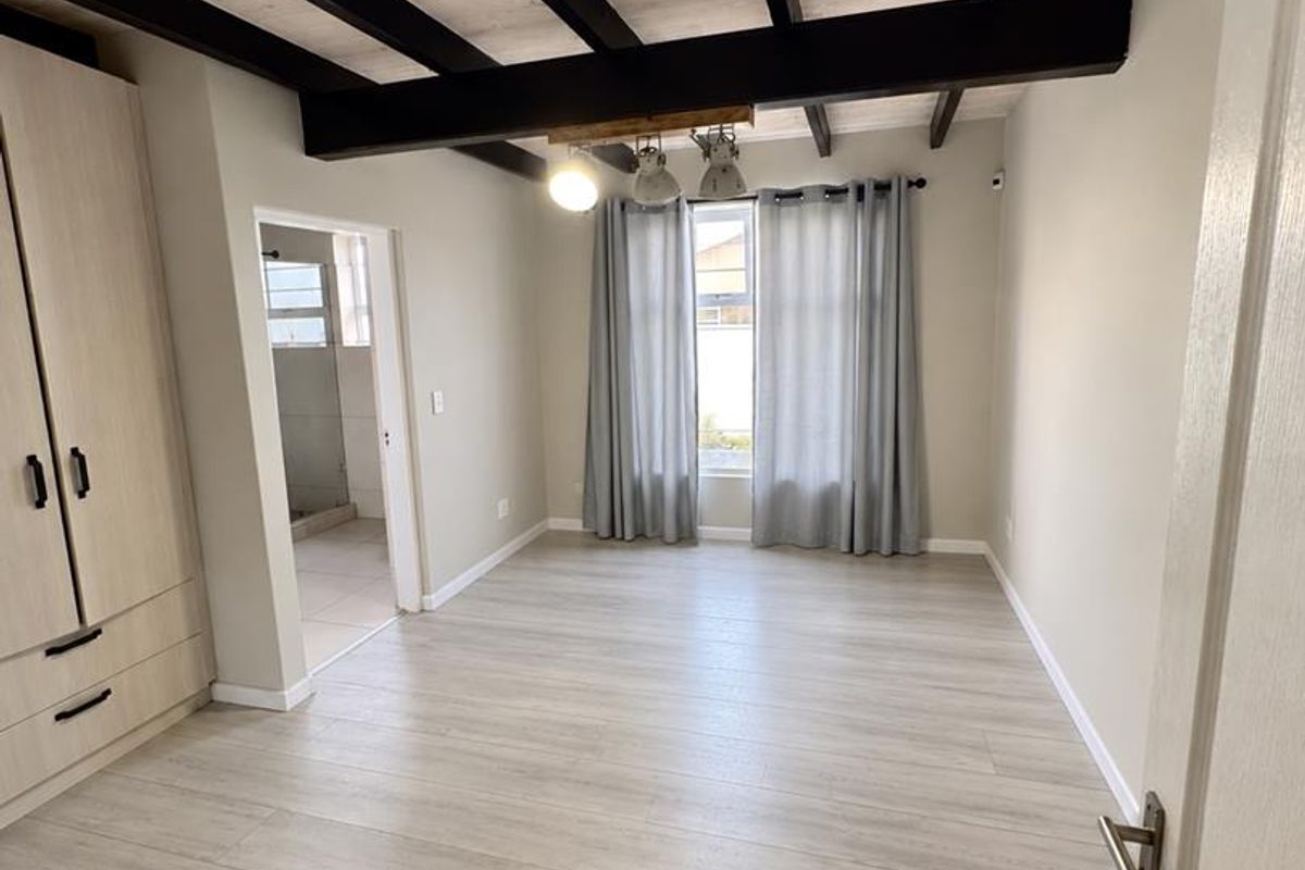 A bright, empty bedroom featuring exposed timber ceiling beams, light-toned laminate flooring, and a built-in wardrobe with an adjoining en-suite bathroom.