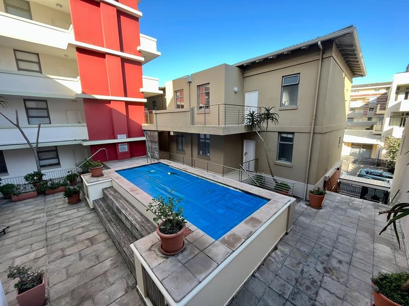 A communal outdoor pool area featuring a paved deck, metal safety railings, and access to the surrounding apartment building complex.