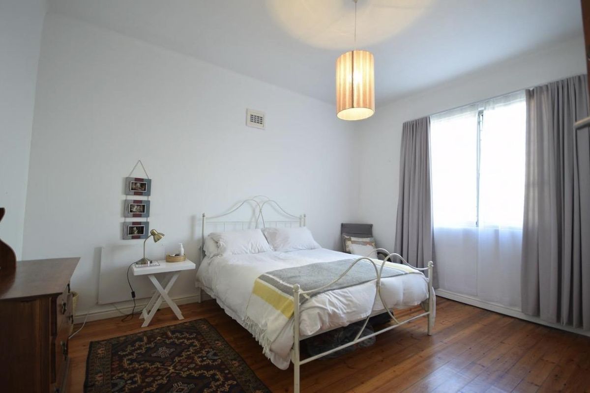 A well-lit bedroom featuring original hardwood flooring, a neutral color palette, and a large window with curtains providing natural light.